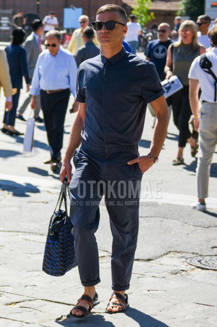 Summer men's coordinate and outfit with plain black sunglasses, plain dark gray jumpsuit, black sports sandals, and black, dark gray, and blue bag briefcase/handbag.