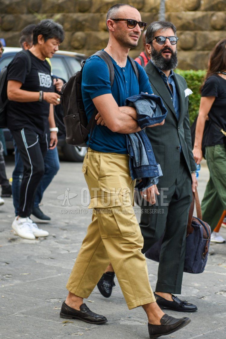 Summer men's coordinate and outfit with brown tortoiseshell sunglasses, plain blue T-shirt with henley neck, plain beige damaged jeans, plain cropped pants, plain cargo pants, black coin loafer leather shoes, and plain brown backpack.