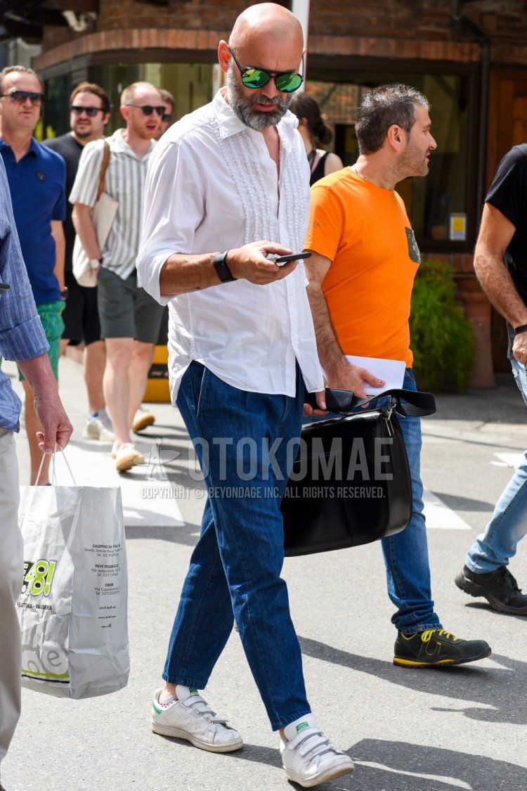 A summer men's outfit and outfit with solid black/green sunglasses, solid white shirt, solid blue denim/jeans, Adidas Stan Smith white low-cut sneakers, solid black shoulder bag, solid black briefcase/handbag.