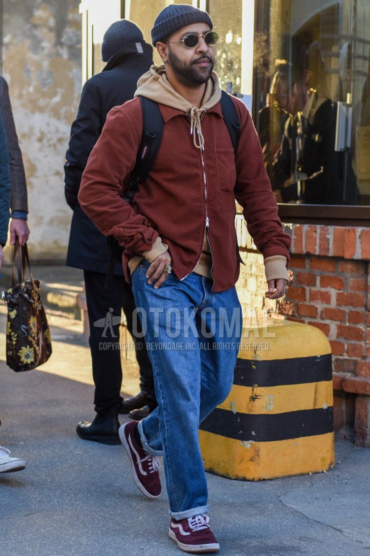Men's fall/winter outfit with solid gray knit cap, solid gold Ray-Ban sunglasses, solid brown shirt jacket, solid beige hoodie, solid blue denim/jeans, and Vans Old Skool red low-cut sneakers.