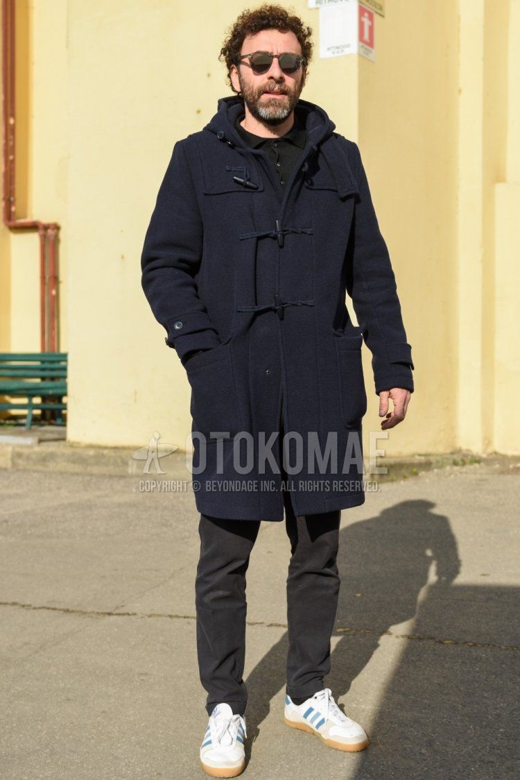 Men's fall/winter outfit with beige tortoiseshell sunglasses, solid navy duffle coat, solid black polo shirt, dark gray solid chinos, solid black socks, and white low-cut Adidas sneakers.
