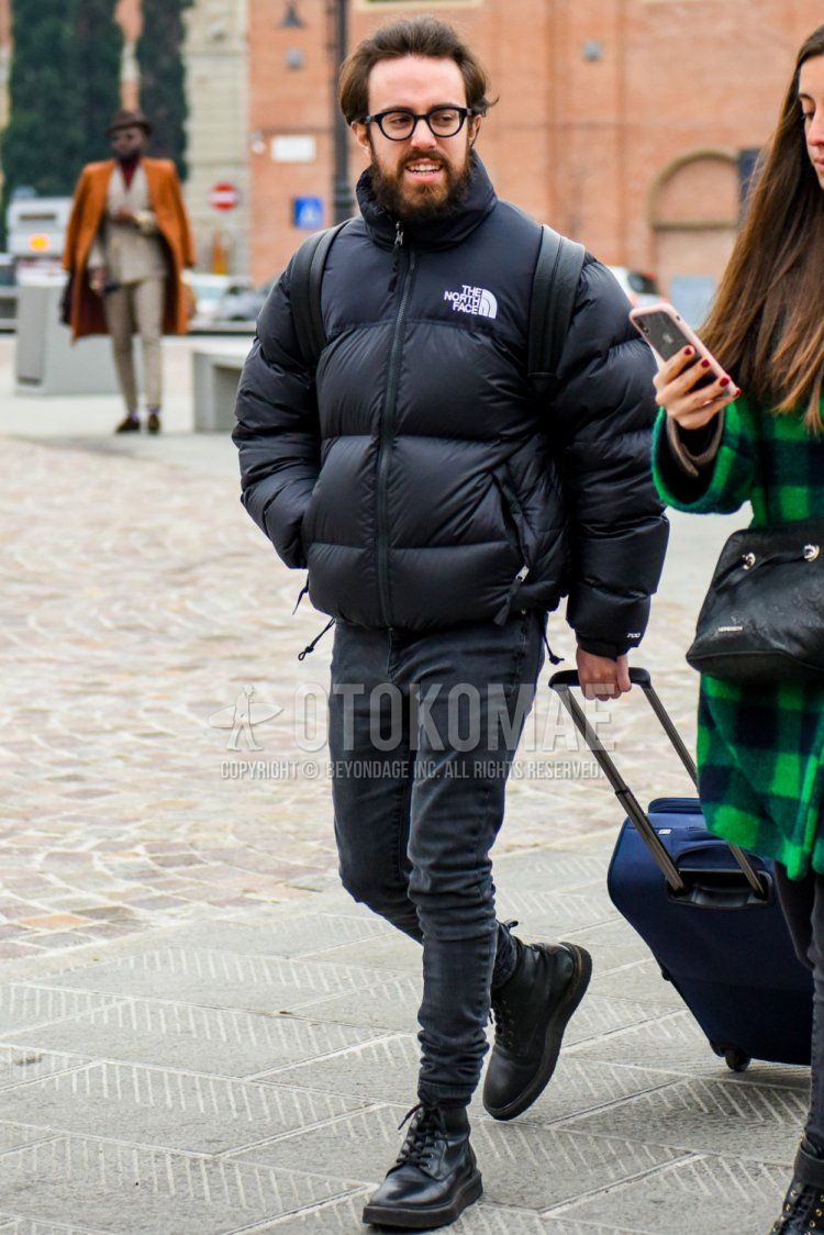 Winter men's coordinate and outfit with plain black glasses, The North Face Nupsi plain black down jacket, dark grey plain denim/jeans, black boots, and a plain grey suitcase.