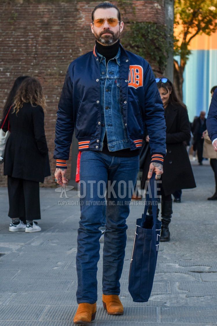 Men's fall/winter outfit with gold solid sunglasses, navy lettered stadium jacket, blue solid denim jacket, black solid turtleneck knit, navy solid denim/jeans, suede brown side gore boots, navy graphic tote bag.