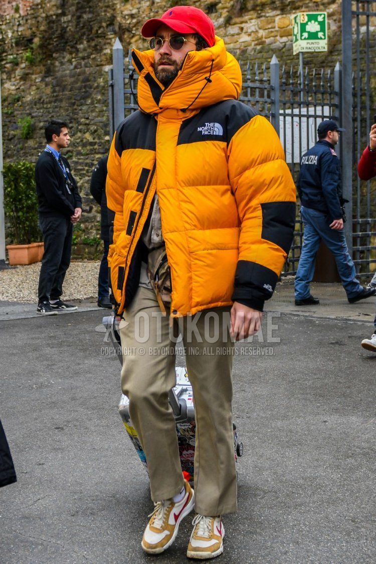 Winter men's coordinate and outfit with red solid color baseball cap, solid color sunglasses, orange solid color down jacket, beige solid color chinos, white solid color socks, and Nike white and brown low-cut sneakers.