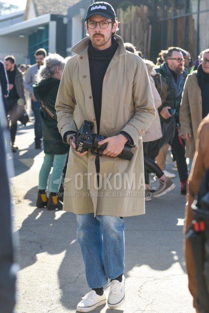 Men's fall/winter coordinate/outfit with black graphic baseball cap, brown tortoiseshell glasses, solid beige stainless steel stainless steel coat, solid dark gray turtleneck knit, solid blue denim/jeans, solid black socks, and white low-cut Vans sneakers.