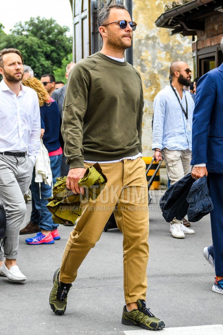 Men's spring and fall coordinate and outfit with brown tortoiseshell sunglasses, plain olive green sweatshirt, plain white t-shirt, plain beige chinos, and Salomon olive green low-cut sneakers.