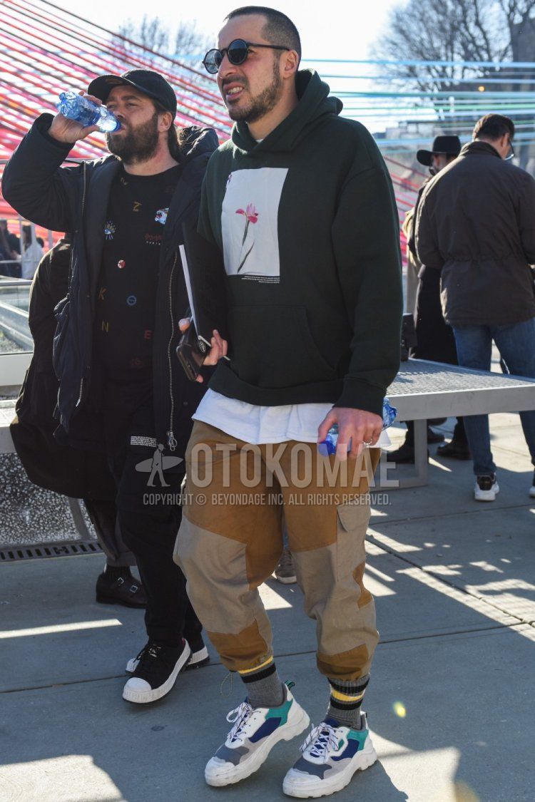 Men's spring/fall outfit with plain black sunglasses, black graphic hoodie, plain white t-shirt, plain brown/beige cargo pants, plain brown/beige jogger pants/ribbed pants, plain gray socks, and gray/white low-cut sneakers.