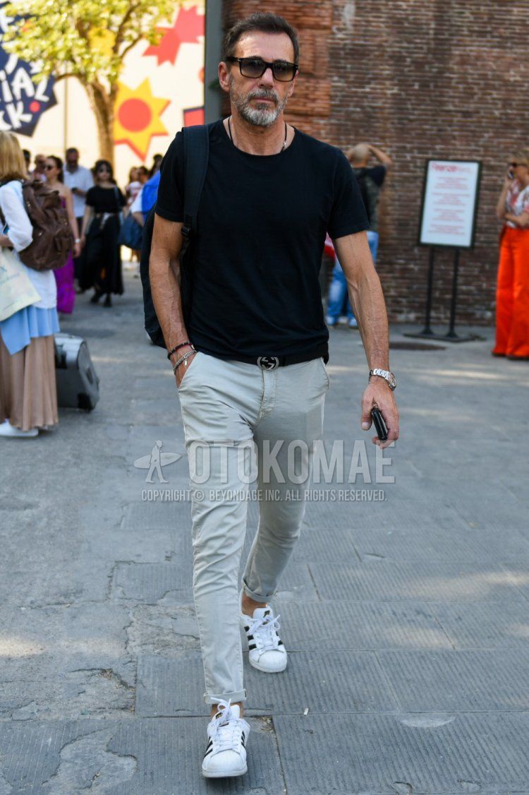 Summer men's coordinate and outfit with plain black sunglasses, plain black t-shirt, Gucci plain black leather belt, plain beige/gray chinos, and Adidas Superstar white low-cut sneakers.