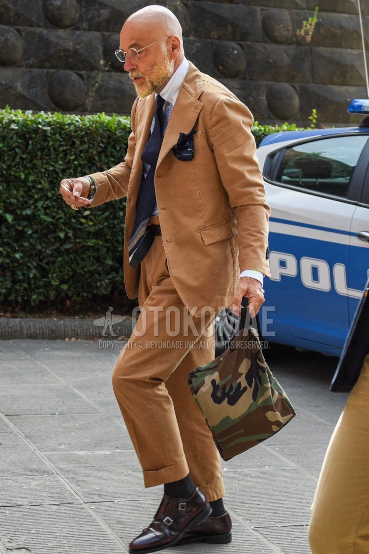 Men's spring/summer/fall coordination and outfit with clear solid glasses, solid white shirt, solid black socks, brown monk shoes leather shoes, multi-colored camouflage bag, solid brown suit, and solid navy tie.