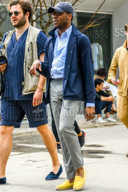 Men's spring, summer, and fall coordinate and outfit with one-pointed baseball cap, solid color sunglasses, solid color navy coach jacket, solid color blue shirt, solid color gray skinny pants, and solid color yellow espadrilles.