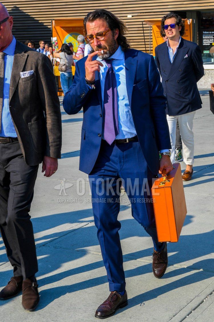Men's spring/summer/fall coordination and outfit with tortoiseshell glasses, light blue/white striped shirt, brown brogue shoes leather shoes, solid orange briefcase/handbag, and solid navy suit.