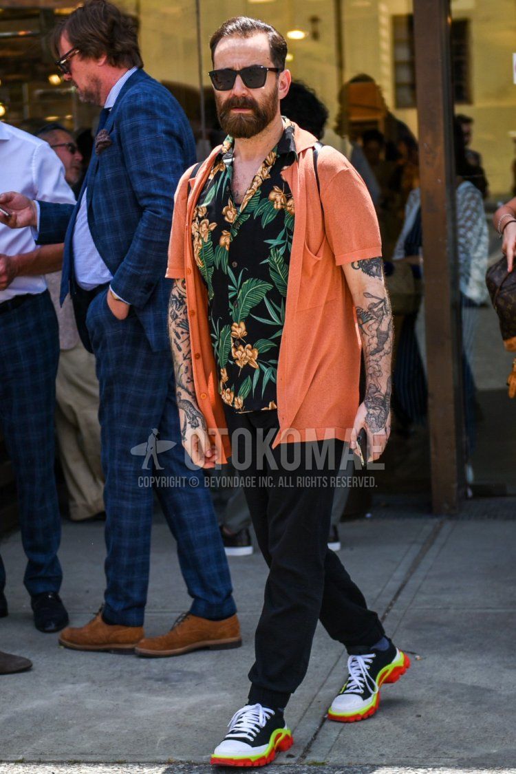 Summer men's coordinate and outfit with solid black sunglasses, solid orange cardigan, multi-colored botanical shirt, solid black jogger pants/ribbed pants, and multi-colored low-cut sneakers.