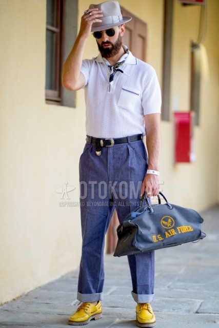 Spring/summer men's outfit with plain gray hat, plain gold sunglasses, white scarf, plain white polo shirt, plain black leather belt, plain blue/navy denim/jeans, yellow moccasin/deck shoes leather shoes, and navy graphic Boston bag. Outfit.