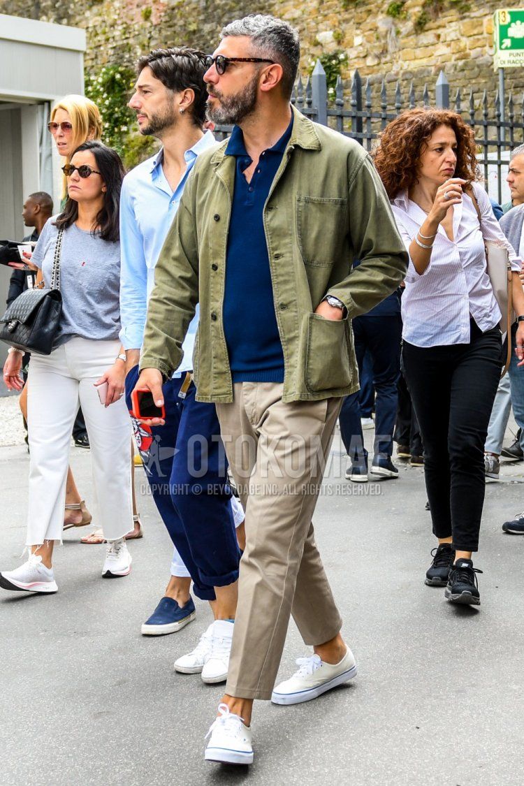Men's spring, summer, and fall coordinate and outfit with solid color sunglasses, olive green solid color shirt jacket, navy solid color polo shirt, beige solid color ankle pants, and white low-cut sneakers.