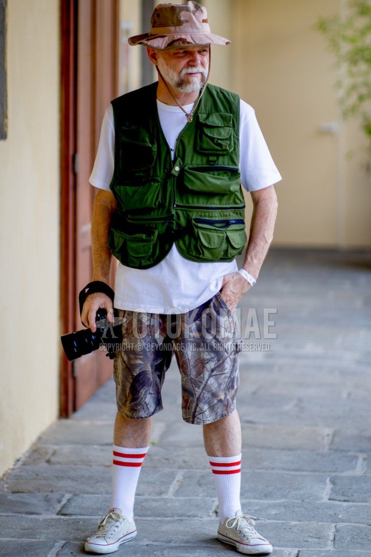 Men's spring and summer coordinate and outfit with beige camouflage cap, olive green solid color casual vest, white solid color t-shirt, brown camouflage shorts, white border socks, and white Converse sneakers.