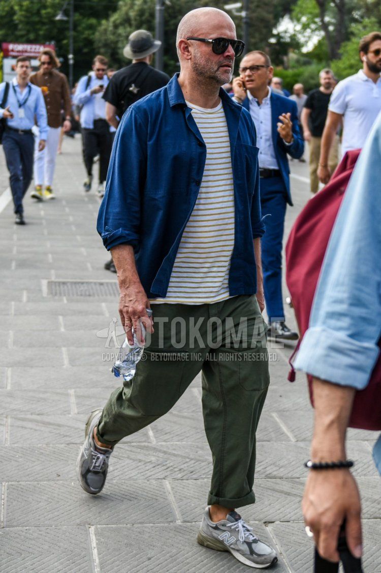Spring and fall men's coordinate and outfit with teardrop solid black sunglasses, solid blue shirt jacket, solid white/yellow t-shirt, solid olive green cargo pants, and New Balance 991 gray low-cut sneakers.