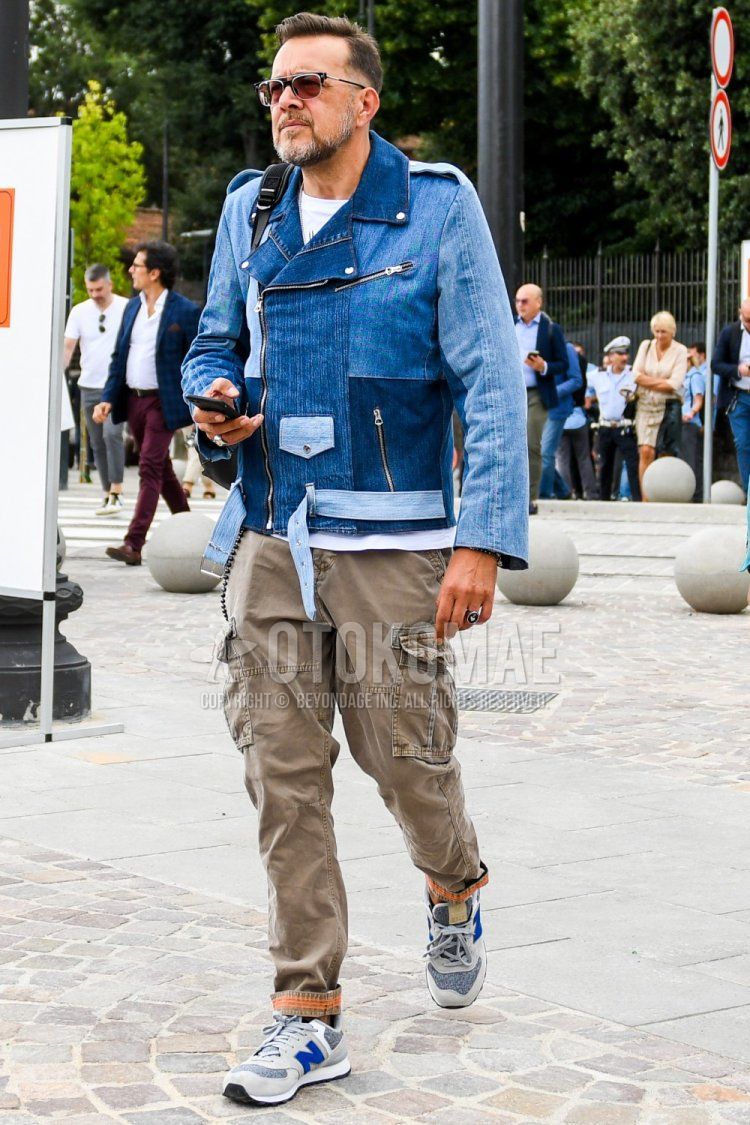 Men's spring, summer, and fall coordinate and outfit with solid color sunglasses, solid color blue rider's jacket, solid color blue denim jacket, solid color white t-shirt, solid color brown cargo pants, and New Balance beige low-cut sneakers.