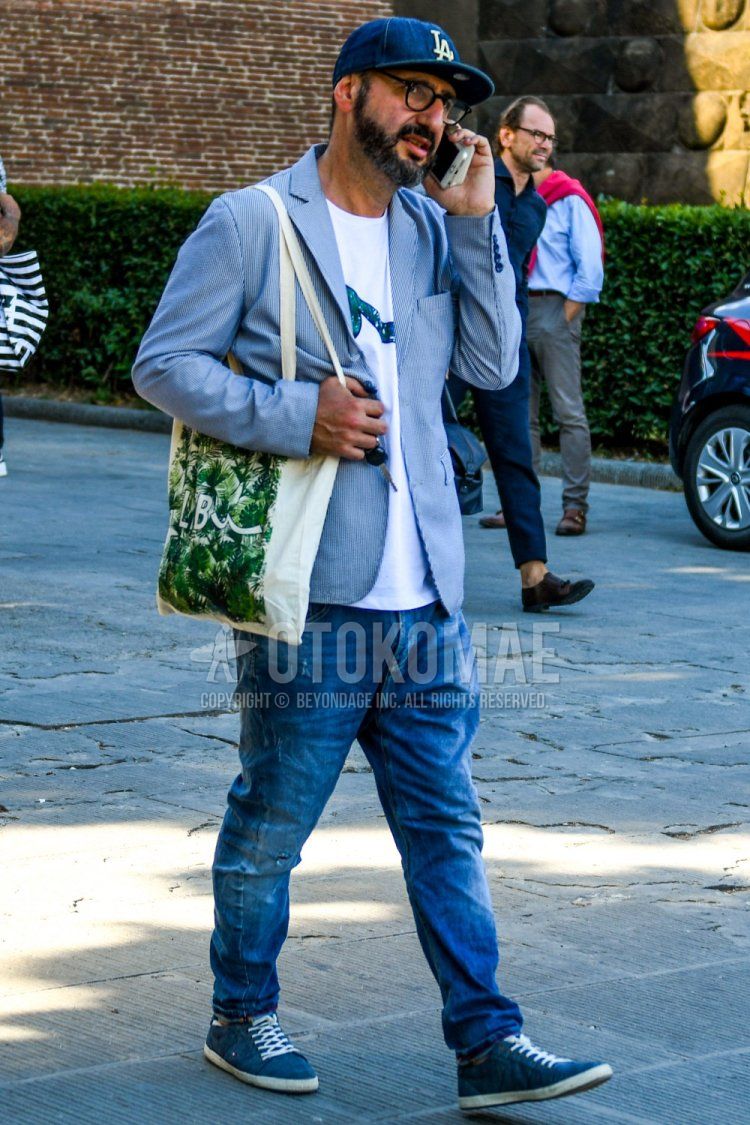Men's spring/summer/fall outfit with plain baseball cap, plain glasses, light blue/white striped tailored jacket, white graphic t-shirt, plain blue denim/jeans, blue low-cut sneakers, and beige graphic tote bag.
