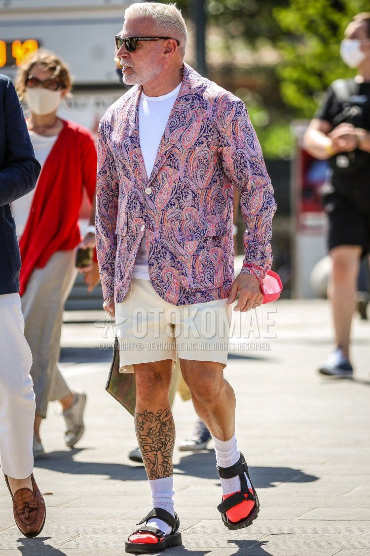 Men's spring/summer coordinate and outfit with brown tortoiseshell sunglasses, purple paisley tailored jacket, plain white t-shirt, plain beige/white shorts, plain white socks, and black sports sandals.