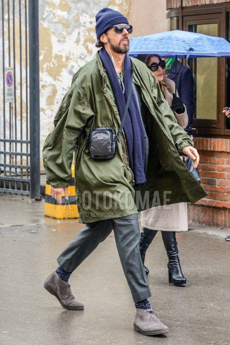 Men's fall/winter outfit and outfit with solid color knit cap, solid color sunglasses, solid color gray scarf/stall, solid color olive green outerwear, solid color gray slacks, black socks, gray chukka boots, and solid color black Supreme body bag.