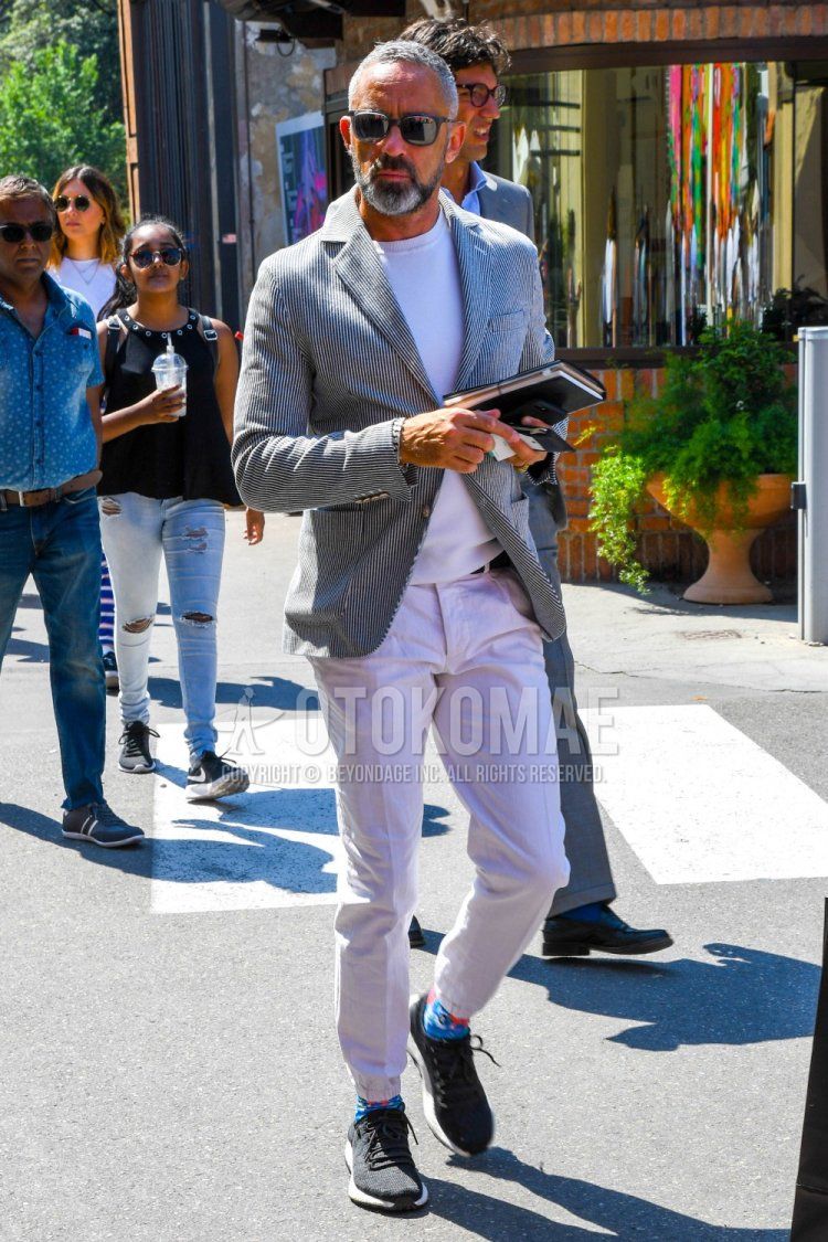 Men's spring/summer/fall outfit with plain sunglasses, black/white striped tailored jacket, plain white sweater, plain white jogger pants/ribbed pants, blue graphic socks, and dark gray low-cut sneakers.