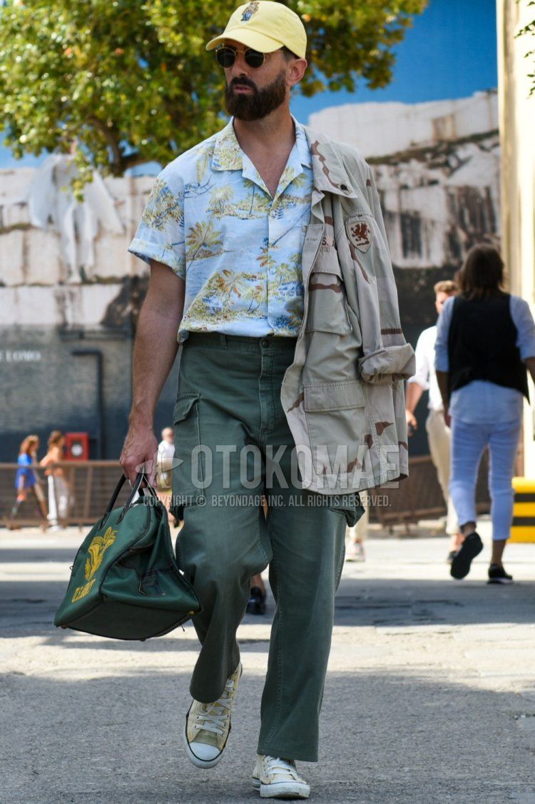Men's spring/summer coordinate and outfit with solid yellow baseball cap, solid gold sunglasses, beige camouflage shirt jacket, light blue graphic shirt, solid olive green cargo pants, beige high-cut sneakers, and solid green Boston bag.