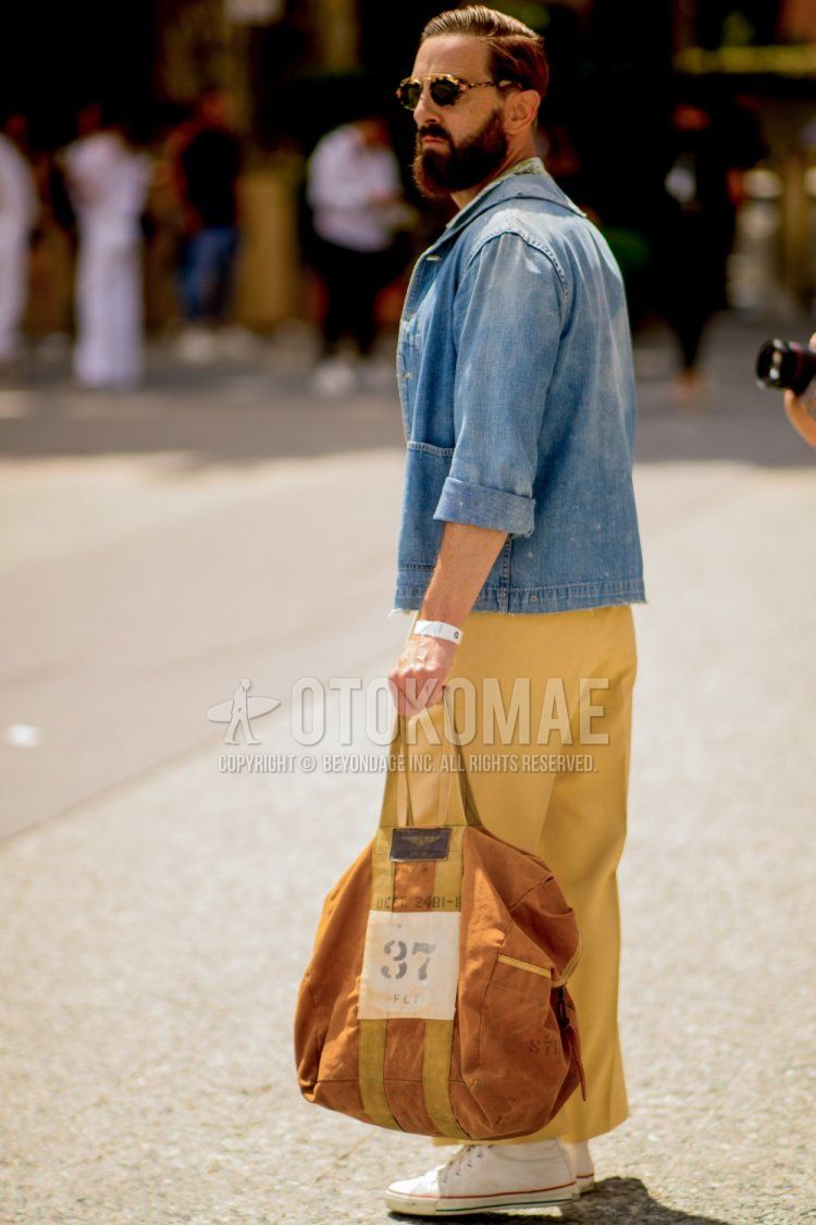 Men's spring/summer outfit with brown tortoiseshell sunglasses, light blue/blue solid denim jacket, green botanical shirt, yellow solid cropped pants, white Converse sneakers, and orange/brown solid tote bag.