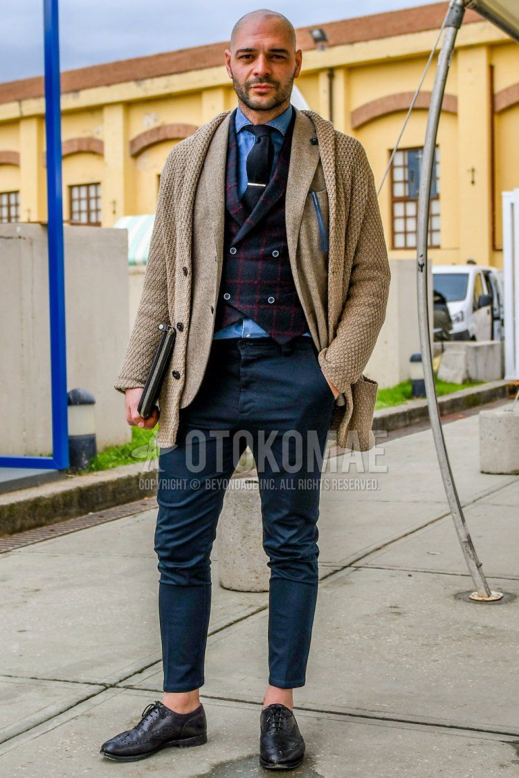 Men's fall/winter outfit with solid beige cardigan, solid beige tailored jacket, gray/red checked gilet, solid blue denim/chambray shirt, solid gray slacks, black wingtip leather shoes, and solid black tie.