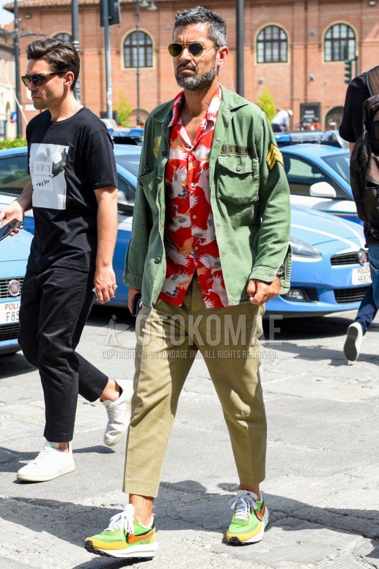 Men's spring/summer/fall outfit with teardrop gold solid color sunglasses, green solid color safari jacket, open collar red top/inner shirt, beige solid color ankle pants, and Nike Sakai green low cut sneakers.