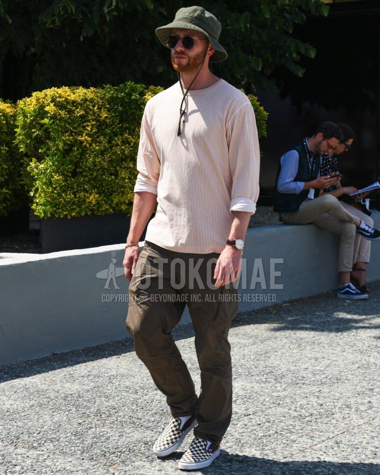 Men's spring/summer coordinate and outfit with olive green solid bucket hat, solid black sunglasses, orange/white striped long tee, olive green solid bottoms, and white/black slip-on sneakers.