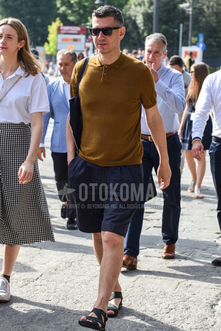 Summer men's coordinate and outfit with square brown tortoiseshell sunglasses, plain brown polo shirt, plain navy shorts, black leather sandals, and plain black tote bag.