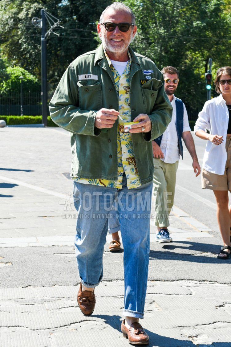 Men's spring/summer coordinate and outfit with plain black sunglasses, plain olive green shirt jacket, plain white t-shirt, yellow botanical shirt, plain blue denim/jeans, and brown tassel loafer leather shoes.