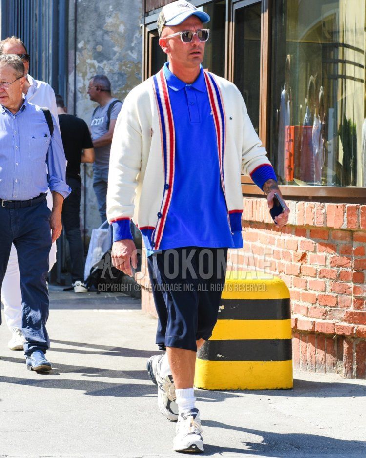Summer/spring men's coordinate and outfit with blue decal logo baseball cap, clear solid color sunglasses, blue solid color polo shirt, beige top/inner cardigan, navy solid color shorts, white solid color socks, white low-cut New Balance sneakers.
