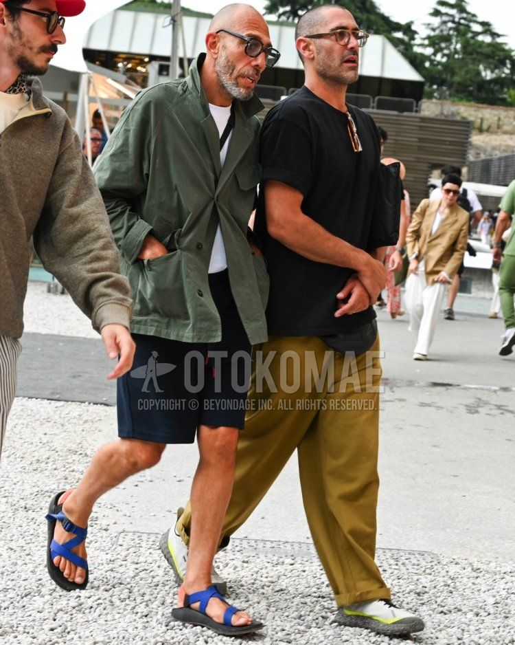Men's spring/summer coordinate and outfit with clear solid color sunglasses, olive green solid color military jacket (other than MA-1 or M-65), white solid color T-shirt, black solid color shorts, and blue sport sandals.