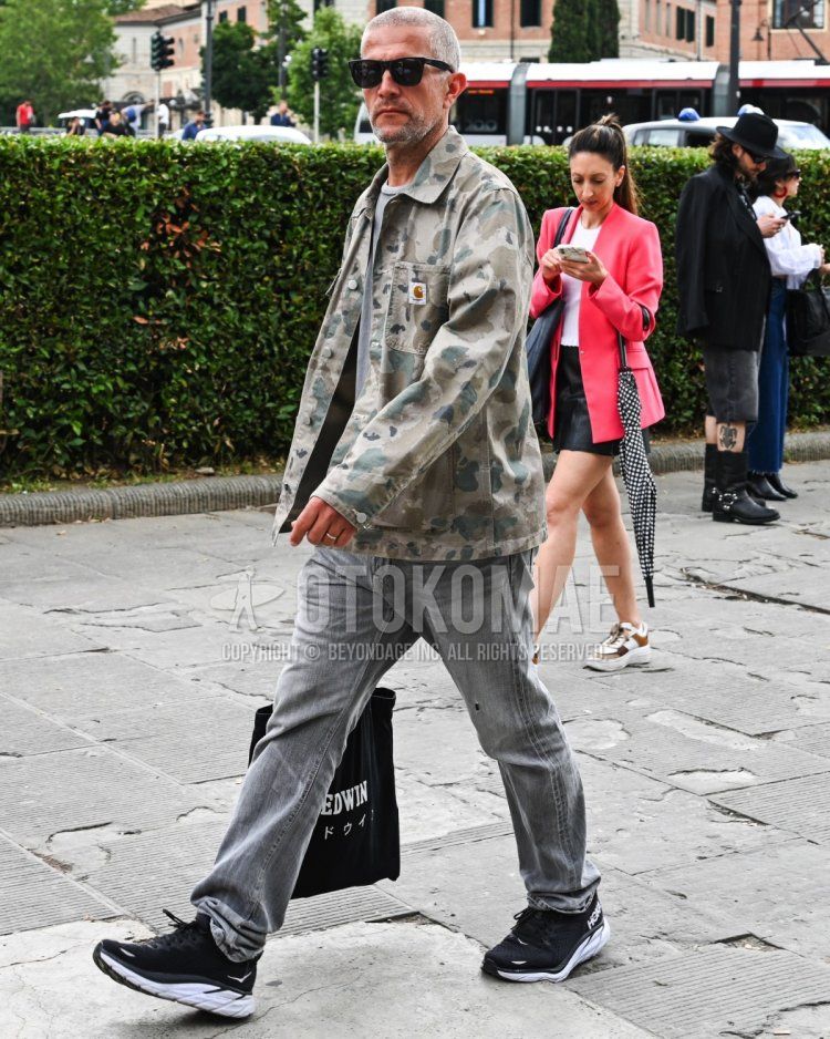 Men's spring/summer/autumn coordinate/outfit with plain black sunglasses, olive green camouflage military jacket (other than MA-1 or M-65), plain gray t-shirt, plain gray/black denim/jeans, and black low-cut sneakers from HOKA.