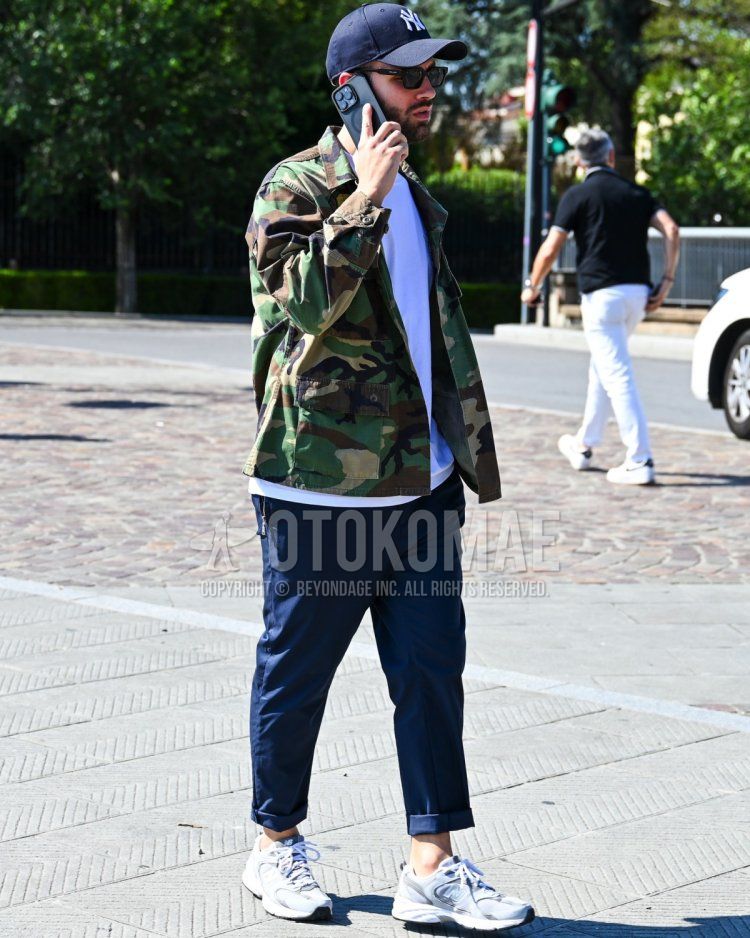 Men's fall/summer/spring coordinate and outfit with navy decal logo baseball cap, plain black sunglasses, olive green camouflage military jacket (other than MA-1 or M-65), plain white t-shirt, plain navy slacks, and white low-cut New Balance sneakers.