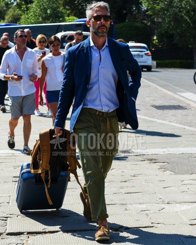 Summer/spring men's coordinate and outfit with brown tortoiseshell sunglasses, plain navy tailored jacket, plain blue shirt, plain olive green bottoms, brown moccasin/deck shoe leather shoes, plain navy suitcase, plain brown backpack.