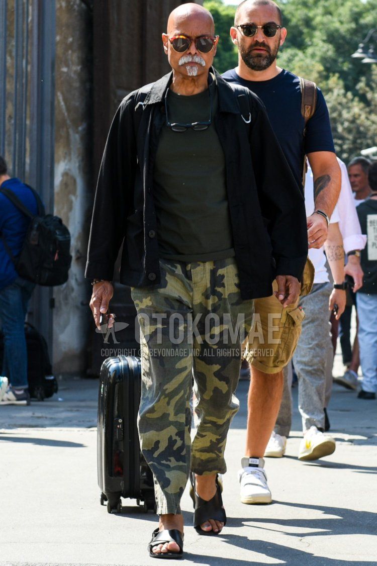 Men's spring/summer coordinate and outfit with brown tortoiseshell sunglasses, plain black shirt jacket, plain olive green t-shirt, olive green camouflage slacks, olive green camouflage ankle pants, and black leather sandals.