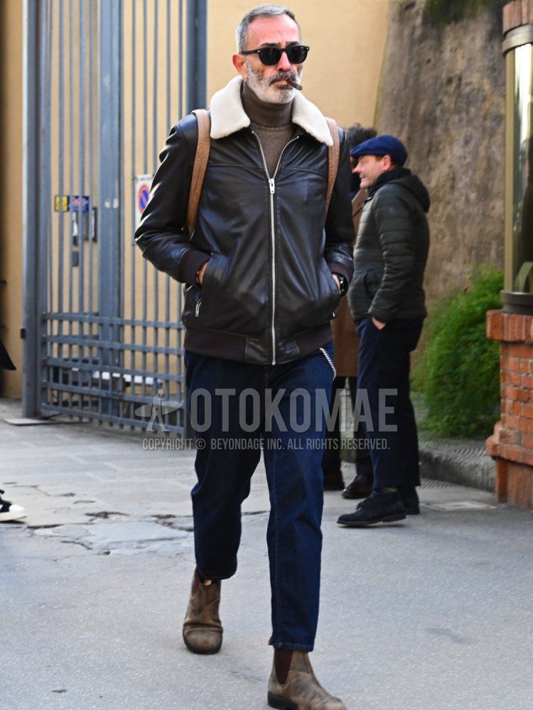Men's fall/winter outfit with solid black sunglasses, solid leather jacket (not rider), solid brown turtleneck knit, solid blue denim/jeans, brown side gore boots, solid brown backpack.