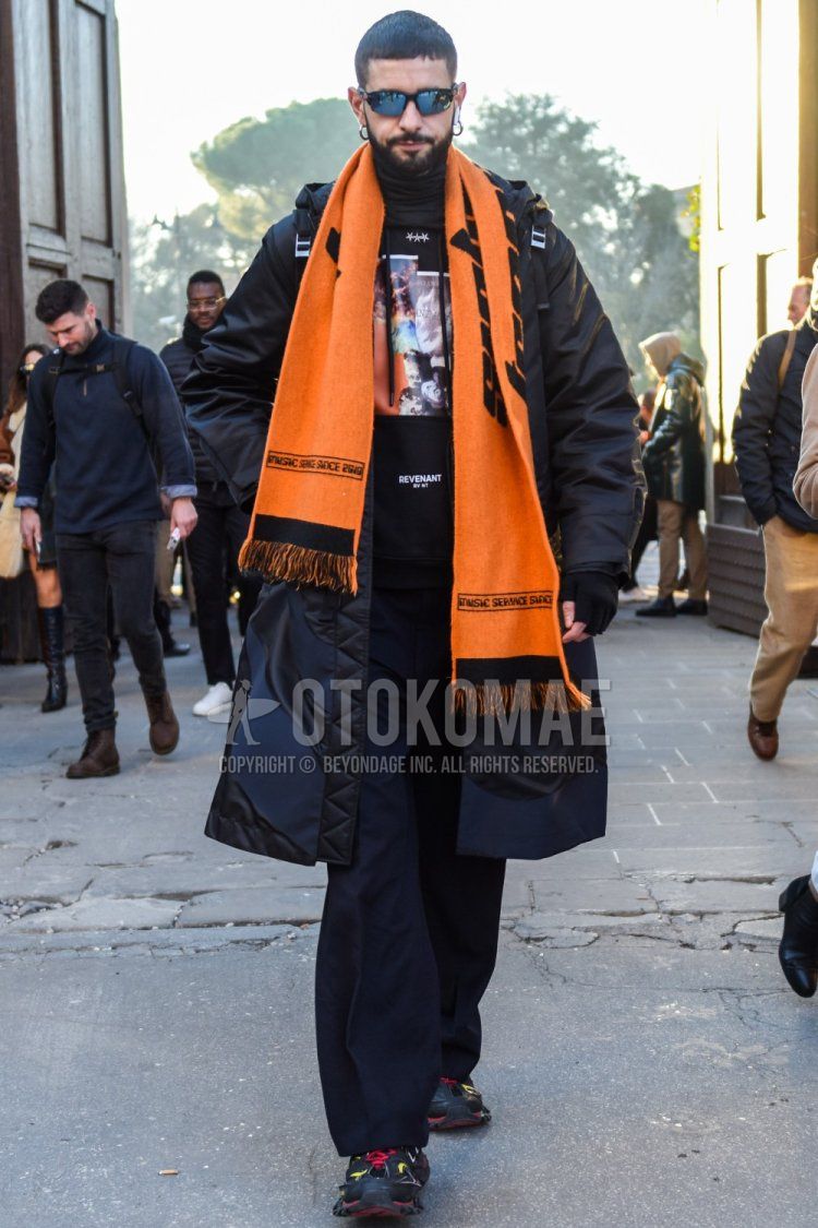 Men's fall/winter outfit with solid black sunglasses, orange graphic scarf/stall, solid black hooded coat, solid black graphic sweatshirt, solid gray slacks, and black low-cut sneakers.