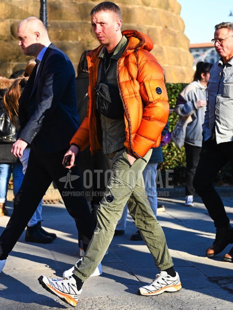 Men's winter/fall outfit with plain orange down jacket, plain olive green coach jacket, plain black t-shirt, plain olive green jogger pants/ribbed pants from cp company, plain black socks, and white low-cut sneakers from Salomon.