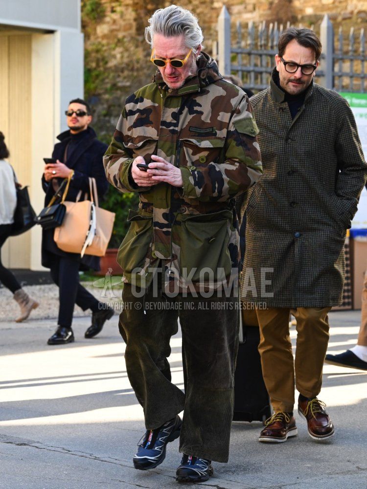 Men's fall/winter outfit with plain yellow sunglasses, olive green camouflage field jacket/hunting jacket, olive green plain winter pants (corduroy,velour), and Salomon navy low-cut sneakers.