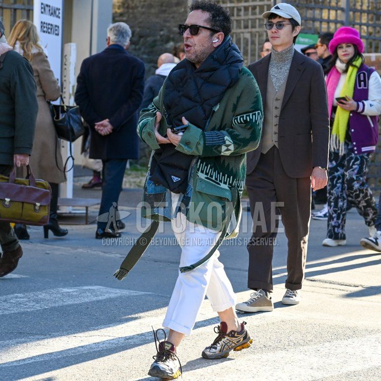 Men's fall/winter outfit with plain black sunglasses, PRADA black one-pointed scarf/stall, olive green all-over pattern outerwear, plain white ankle pants, and asics multi-colored low-cut sneakers.