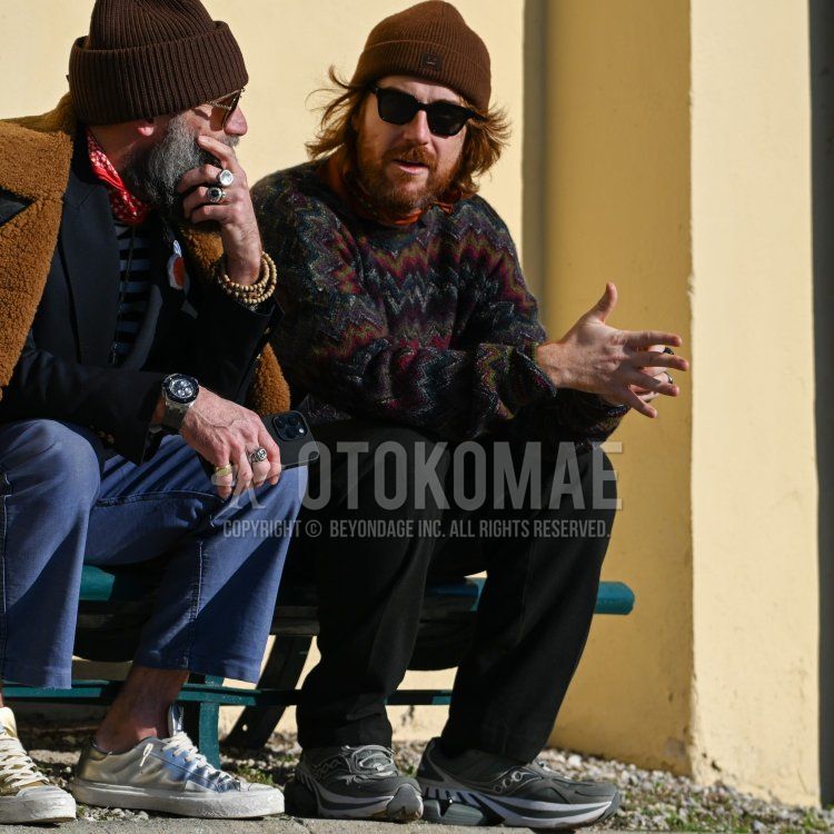 Men's fall/winter coordinate and outfit with plain brown knit cap, plain black sunglasses, plain orange bandana/neckerchief, multi-colored all-over sweater, plain black wide-leg pants, and gray low-cut sneakers.