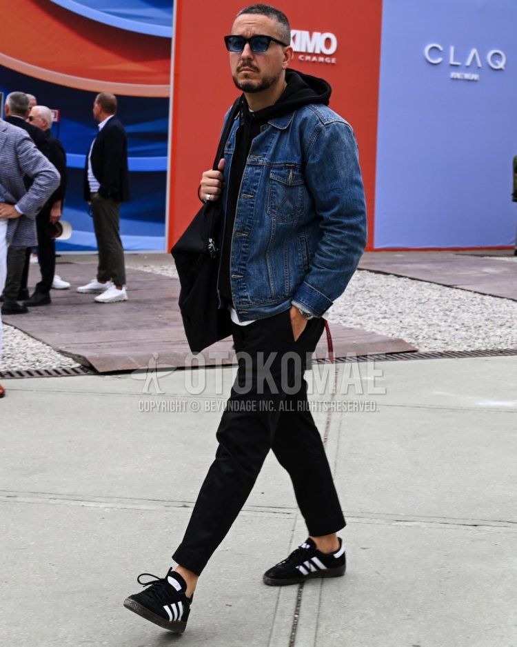 Men's spring and fall outfit with solid blue sunglasses, solid blue denim jacket, solid black hoodie, solid black chinos, and black low-cut Adidas sneakers.