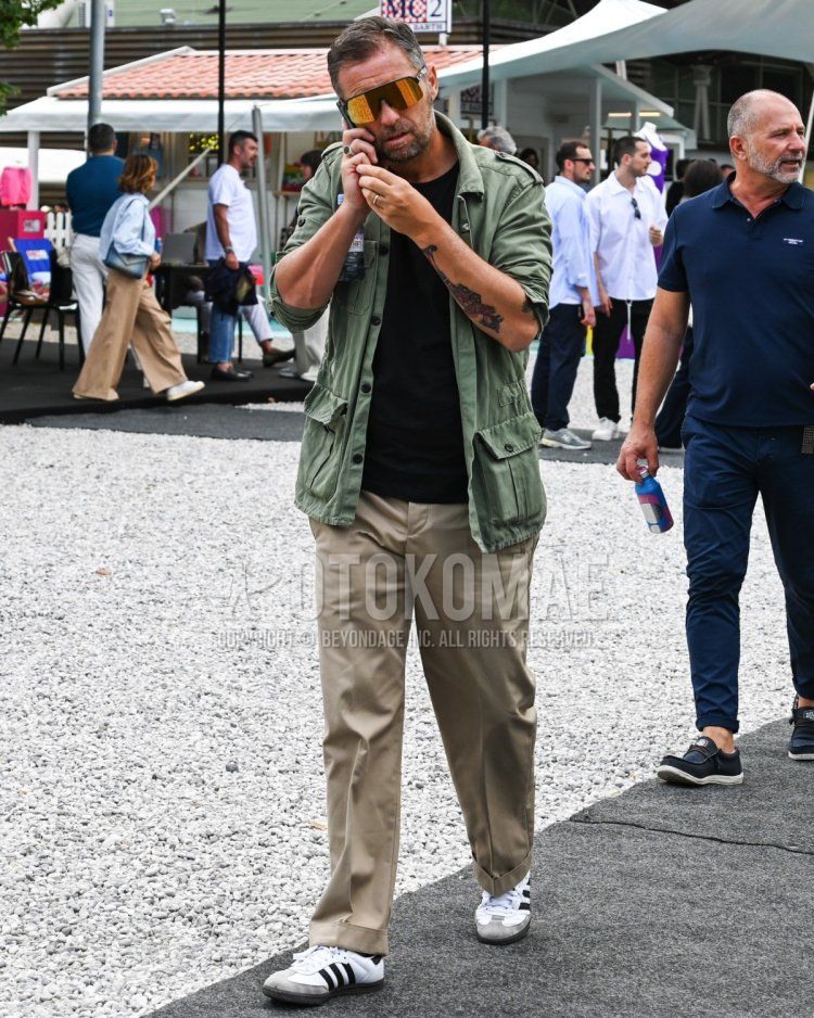 Men's spring, summer, and fall coordinate and outfit with multi-colored solid color sunglasses, olive green solid color M-65, solid color black t-shirt, beige solid color chinos, and white low-cut Adidas sneakers.