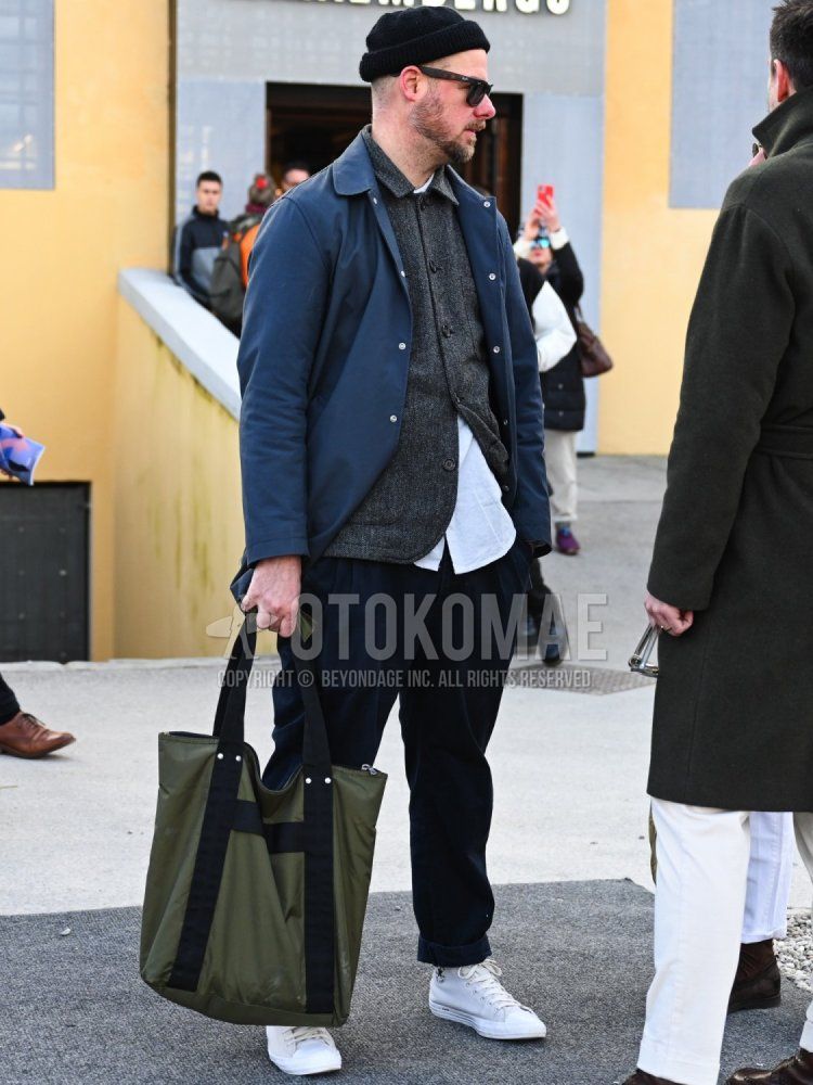 Men's winter/autumn coordinate and outfit with solid black knit cap, solid black sunglasses, solid navy coach jacket, solid gray coveralls, solid white shirt, solid navy cotton pants, white high-cut Converse sneakers, and solid olive green tote bag.