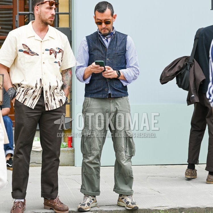 Men's fall/spring outfit with plain black/orange sunglasses, plain navy casual vest, light blue/white striped shirt, plain olive green cargo pants, and New Balance white/beige/brown low-cut sneakers.