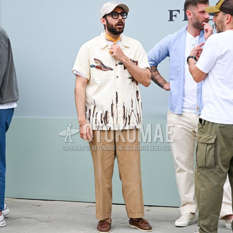 Summer men's coordinate and outfit with solid beige baseball cap, solid black glasses, solid yellow scarf, solid beige, brown, navy, and red graphic shirt, solid beige chinos, and brown moccasin/deck shoe leather shoes.