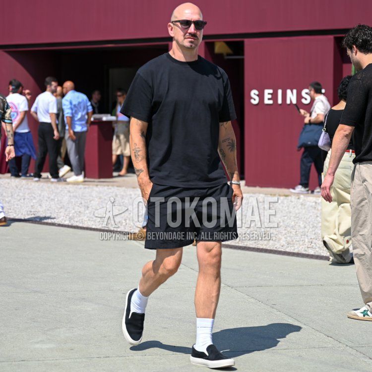 Men's summer coordinate and outfit with brown tortoiseshell sunglasses, plain black t-shirt, plain black shorts, plain white socks, black slip-on sneakers, and black suede shoes leather shoes.